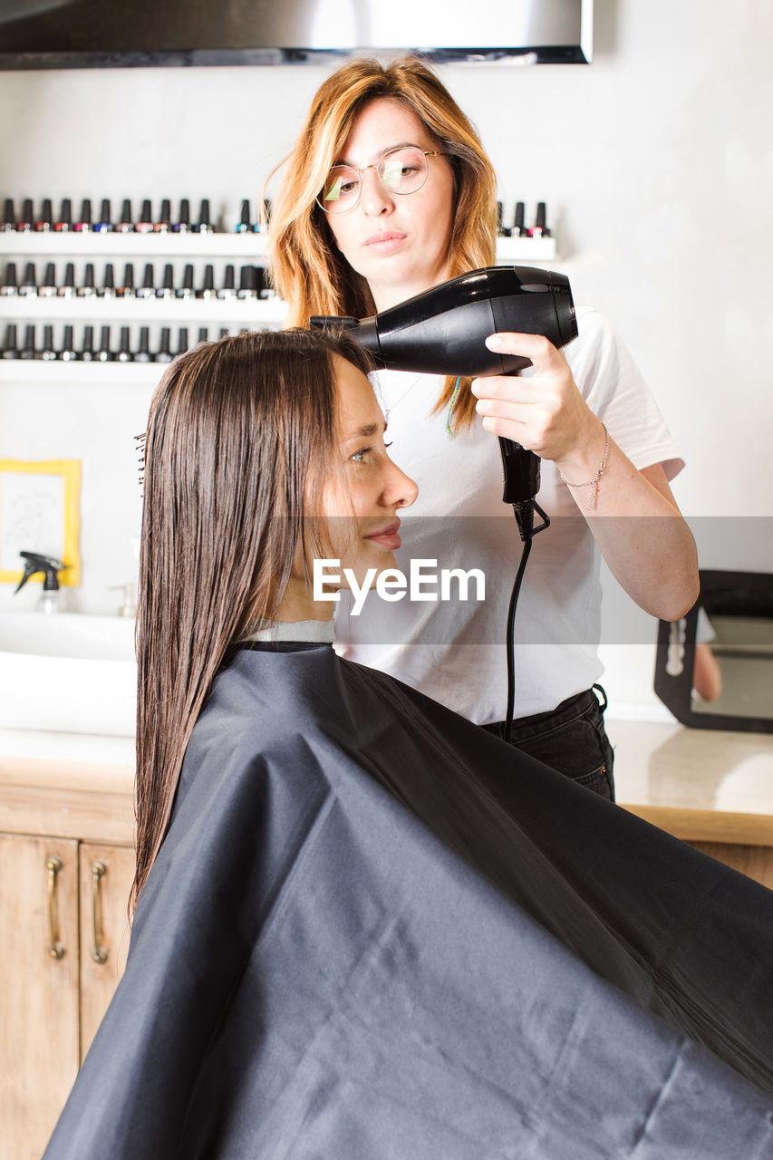 The hairdresser dries the client's hair with a hair dryer and styles her hair with a comb.