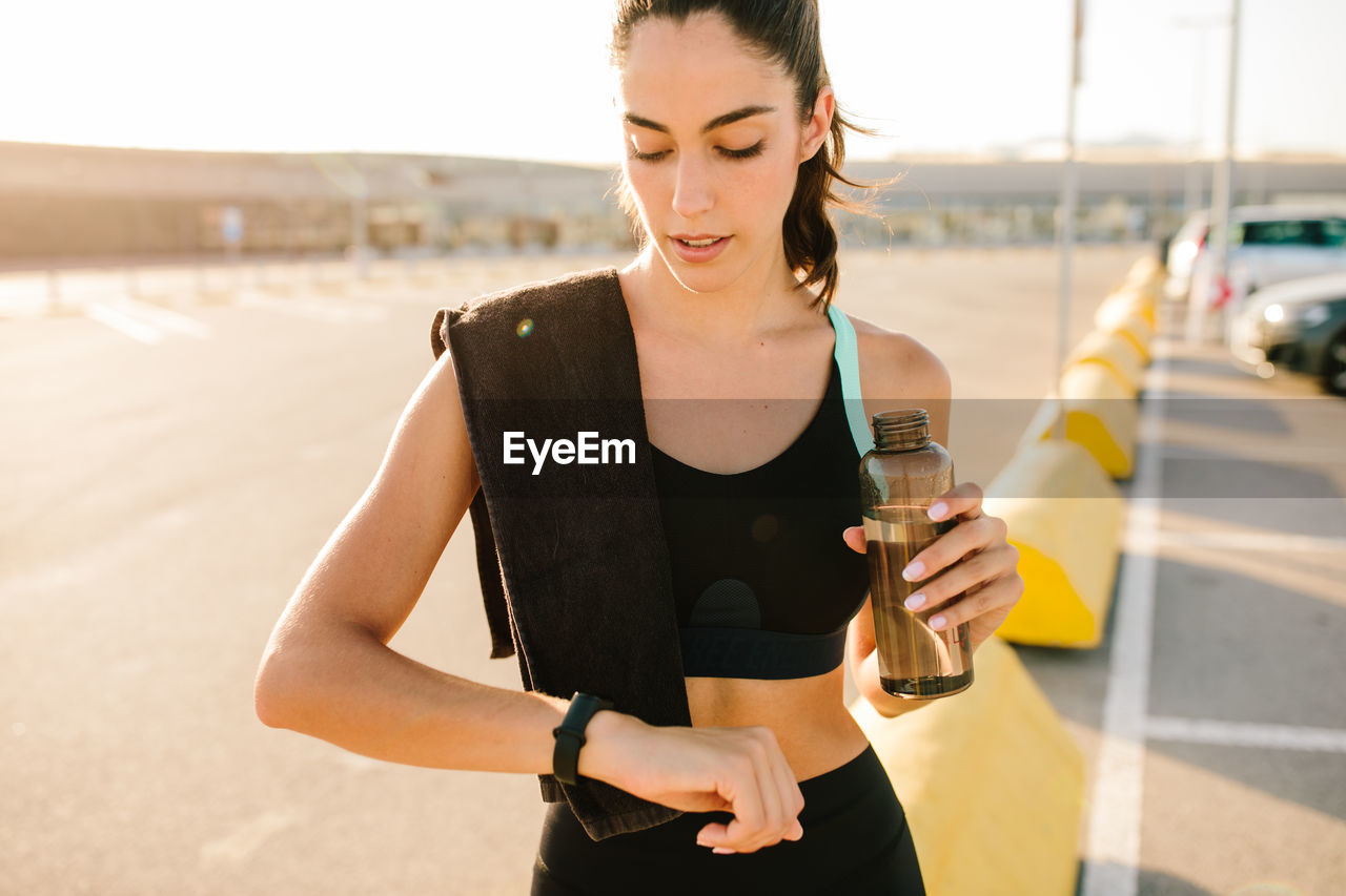 Young fit female in sportswear holding bottle of water and checking sports results on activity tracker while resting after running on street