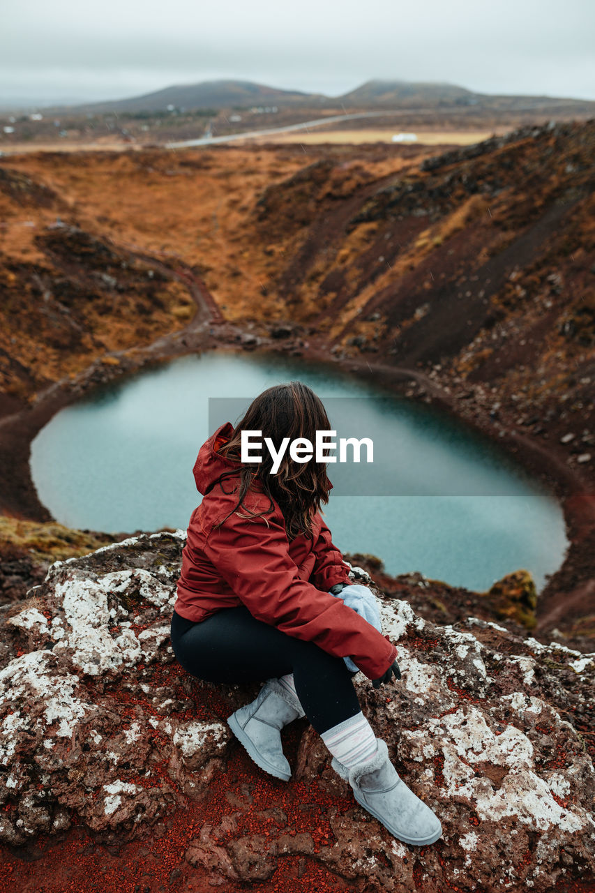 From above side view of young tourist sitting on peak of mountain and looking at water in valley