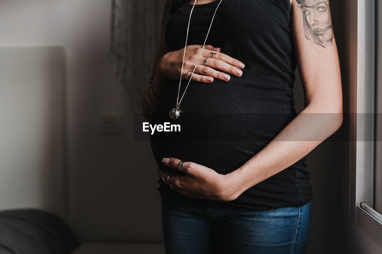 Midsection of pregnant woman standing by window at home