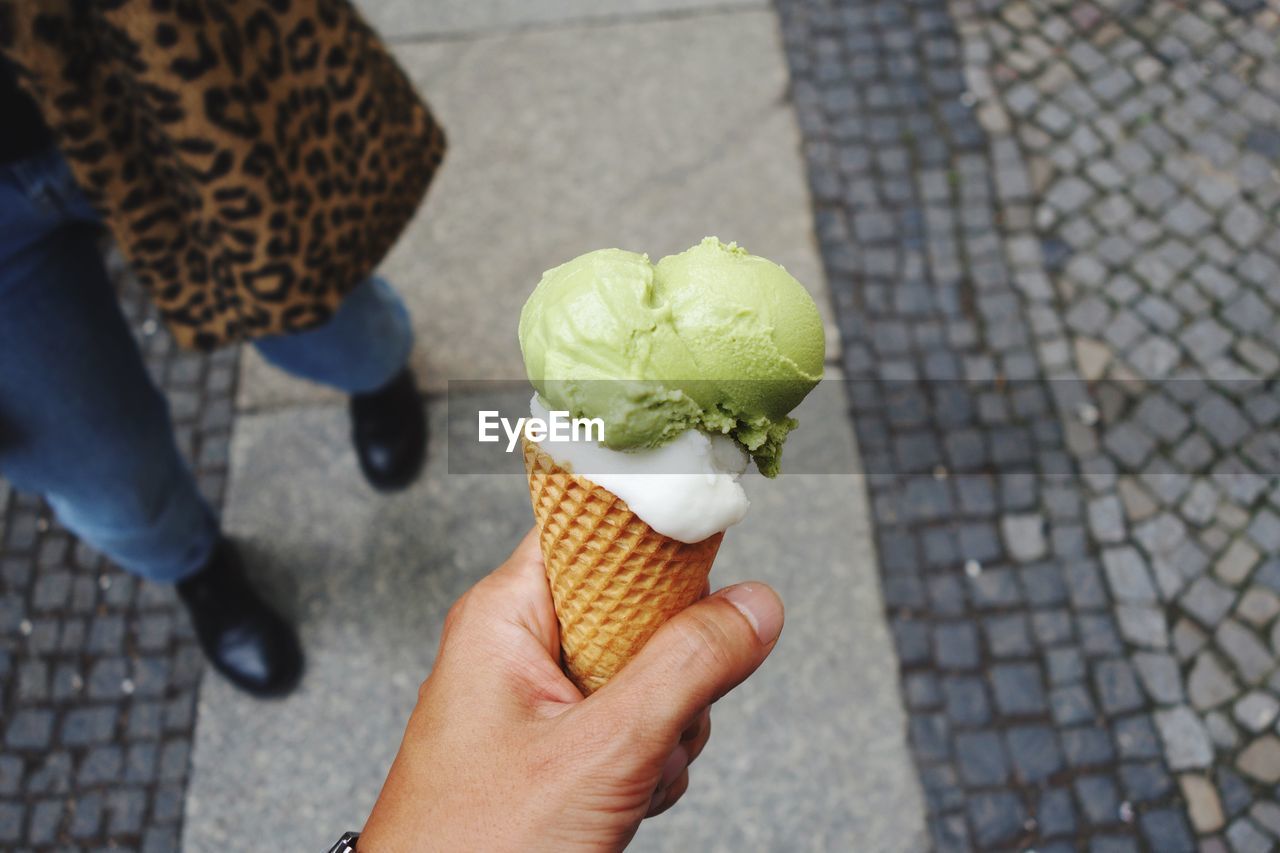 Cropped hand holding ice cream cone by woman standing on footpath