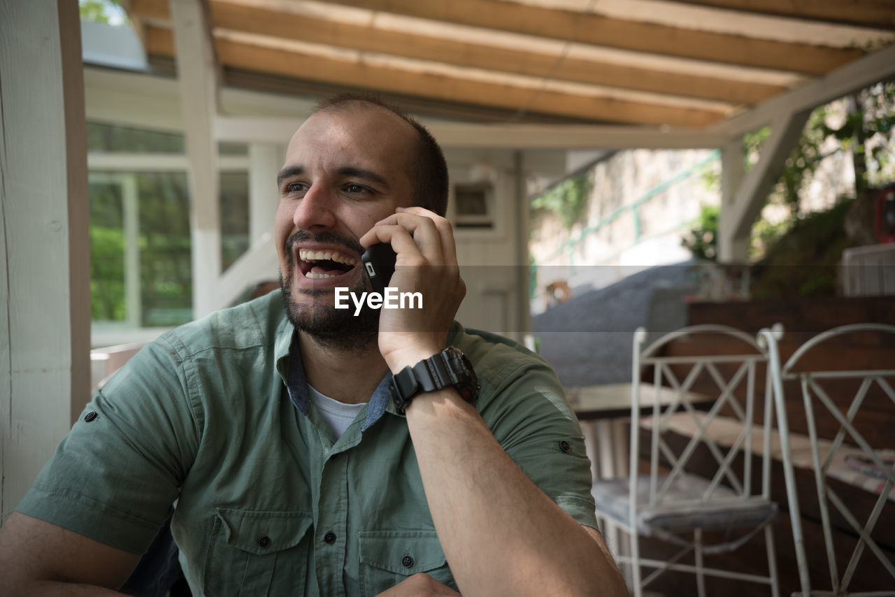 Cheerful young man talking through phone while sitting at yard