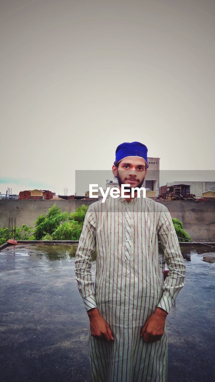 one person, portrait, looking at camera, standing, adult, men, front view, sky, clothing, water, architecture, smiling, three quarter length, nature, emotion, copy space, spring, day, facial hair, person, pride, waist up, happiness, occupation, outdoors, hat, beard, built structure, blue