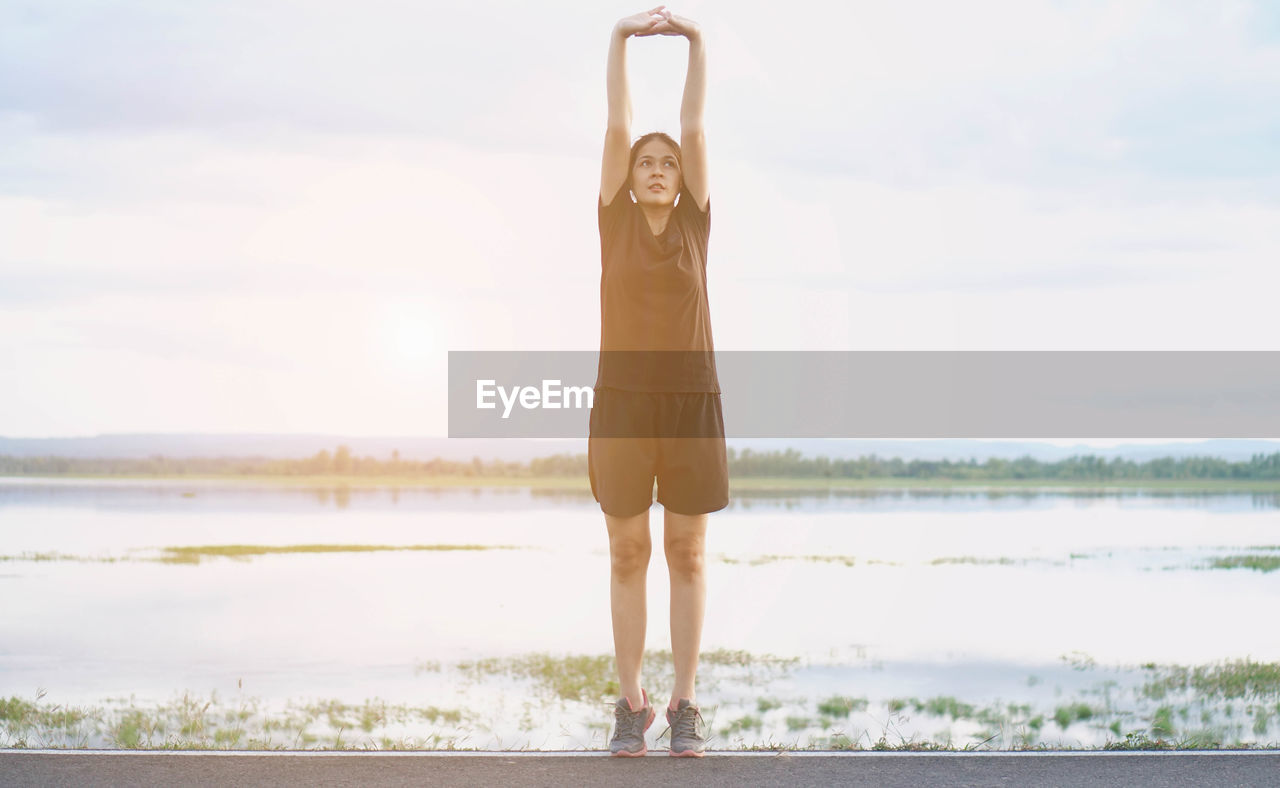 Young woman stretching on road against lake and cloudy sky