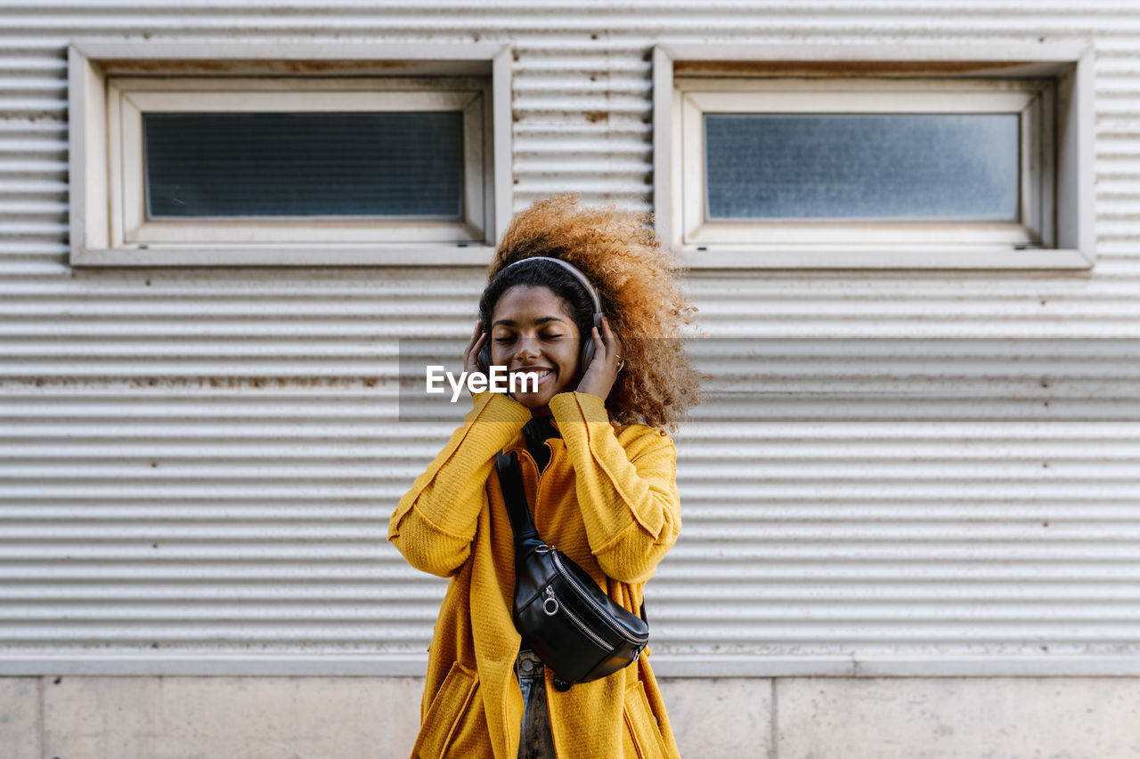 Smiling afro woman with eyes closed listening music through wireless headphones while standing against wall