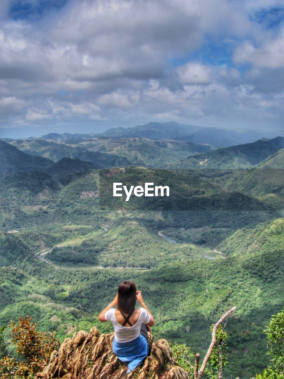 Rear view of mid adult woman looking at view while sitting on mountain against cloudy sky
