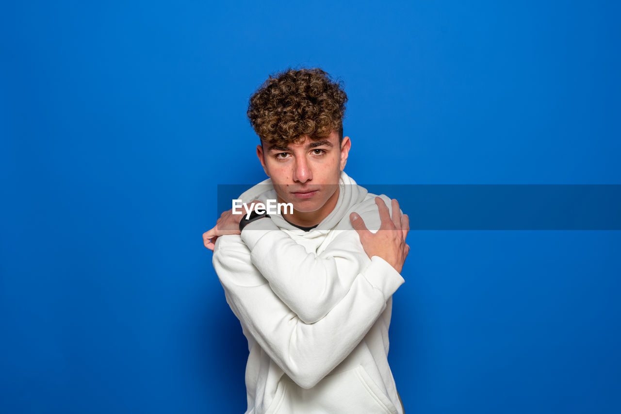 blue, colored background, studio shot, one person, blue background, portrait, adult, person, waist up, young adult, indoors, hand, arm, men, clothing, curly hair, copy space, looking at camera, sitting, human face, front view, finger, emotion, standing, photo shoot, negative emotion