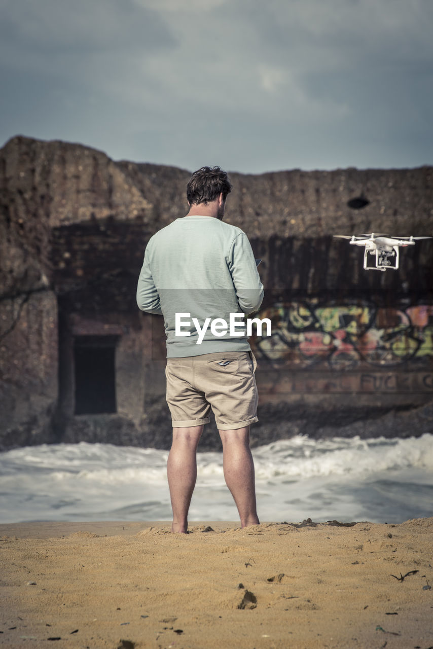 Rear view of man with drone at shore against sky