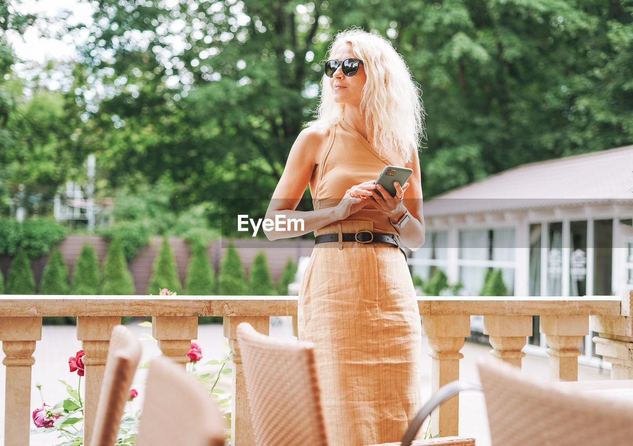 Adult elegant blonde woman in sunglasses and beige clothes using mobile phone in summer cafe