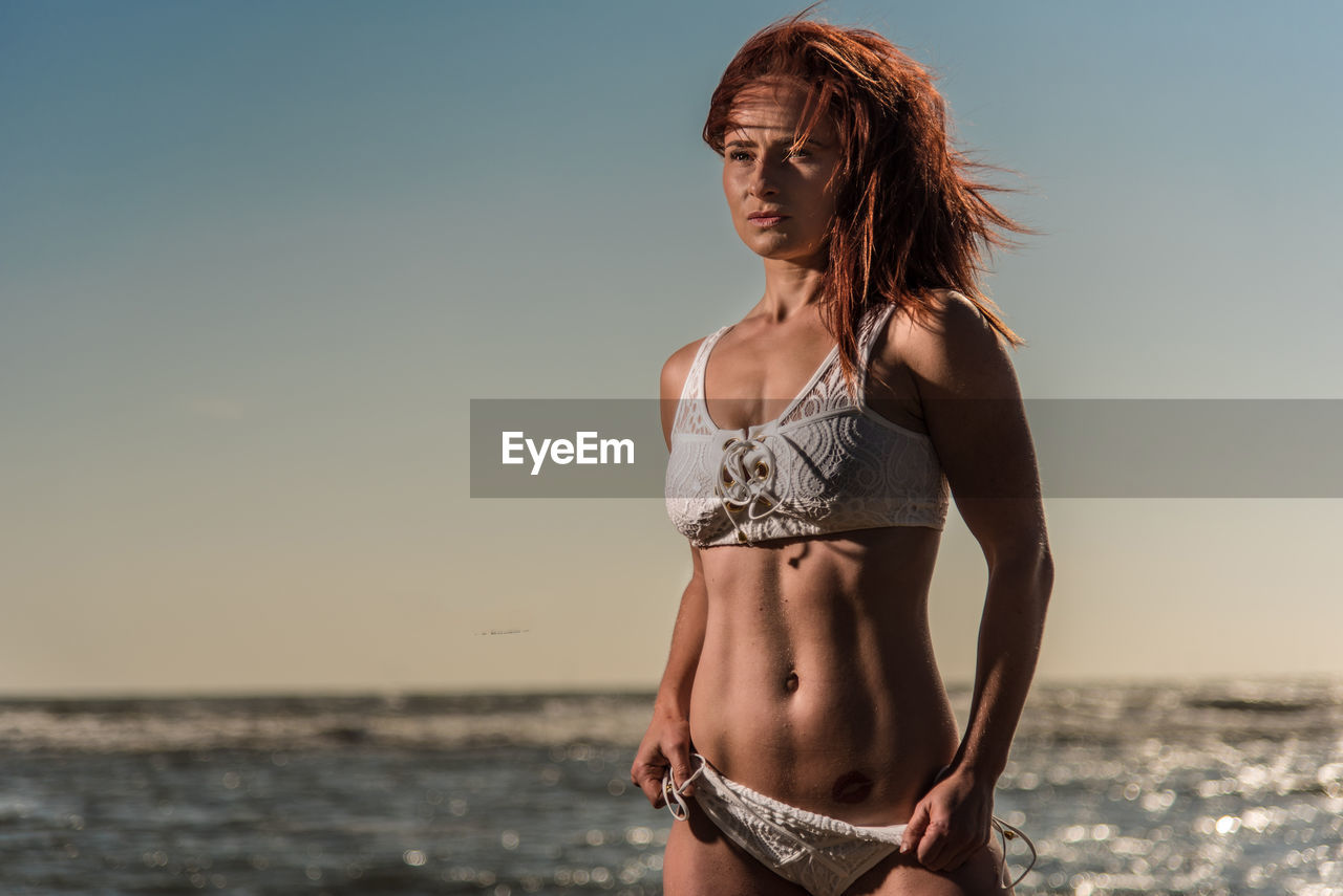 Sensuous female model standing in bikini at beach against sky