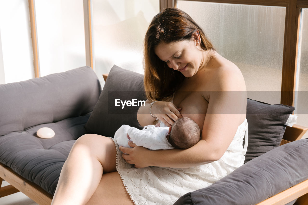 Adult woman smiling and breastfeeding newborn baby while sitting on couch at home