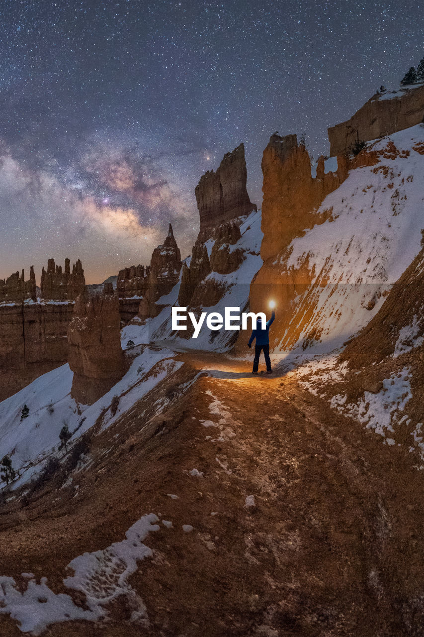 Silhouette of unrecognizable explorer standing with flashlight on scenery of rocky formations in snowy mountains under milky way starry sky in goblin valley state park, utah, usa.