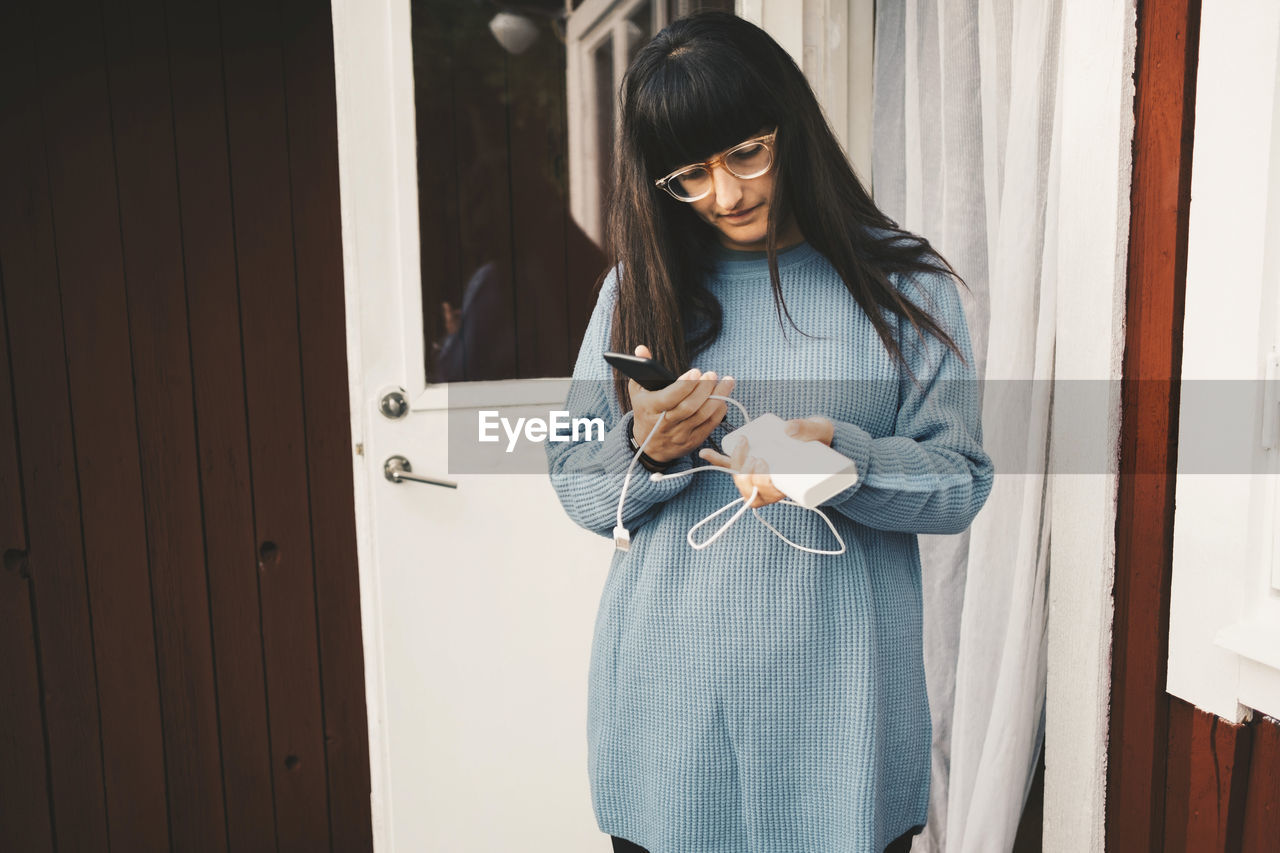 Woman holding smart phone and portable charger while standing against door