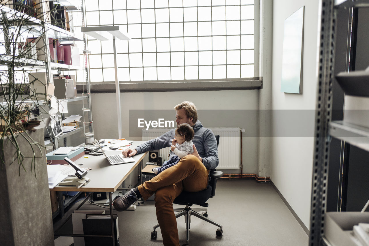 Man with baby on lap using laptop at desk in a factory