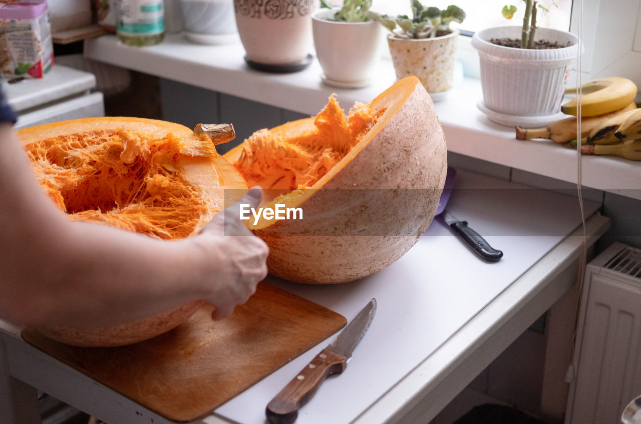 Woman cuts a large pumpkin with a knife on a table by the window