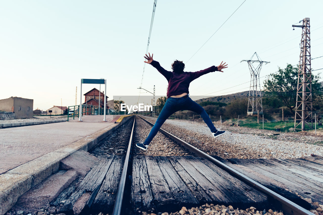 Rear view of teenage girl jumping over railroad tracks against clear sky