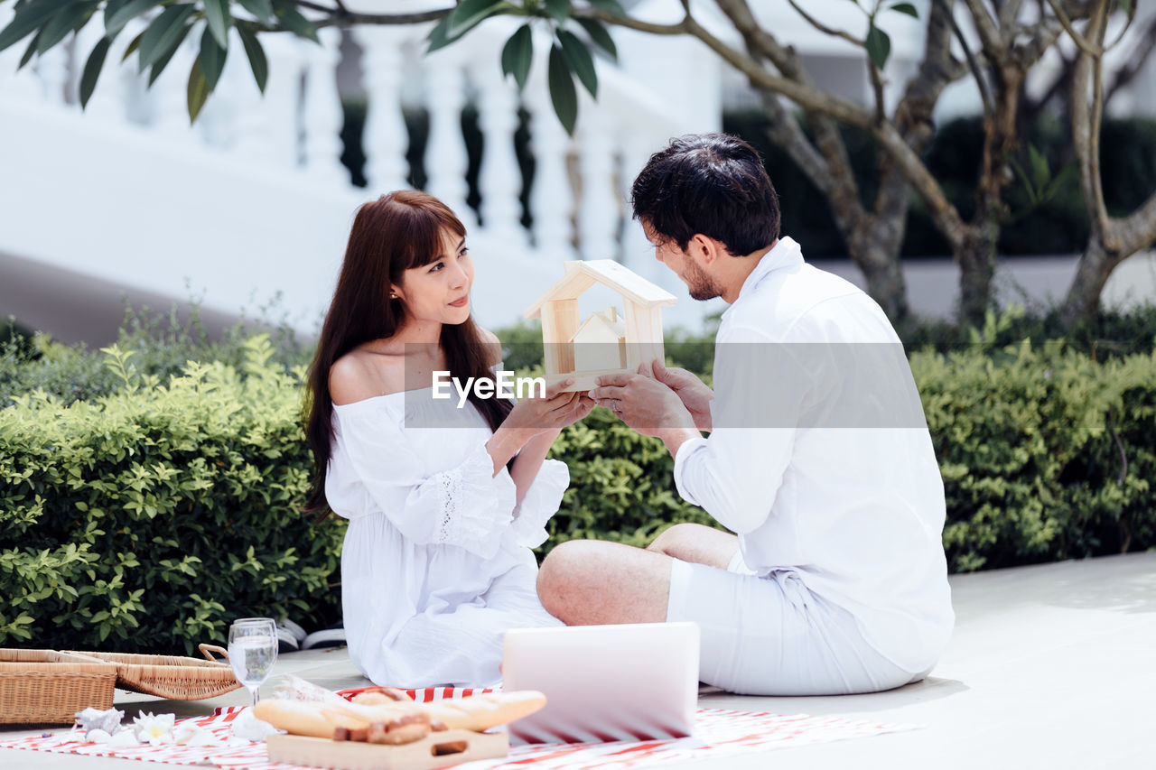 Couple holding model home while sitting by plants