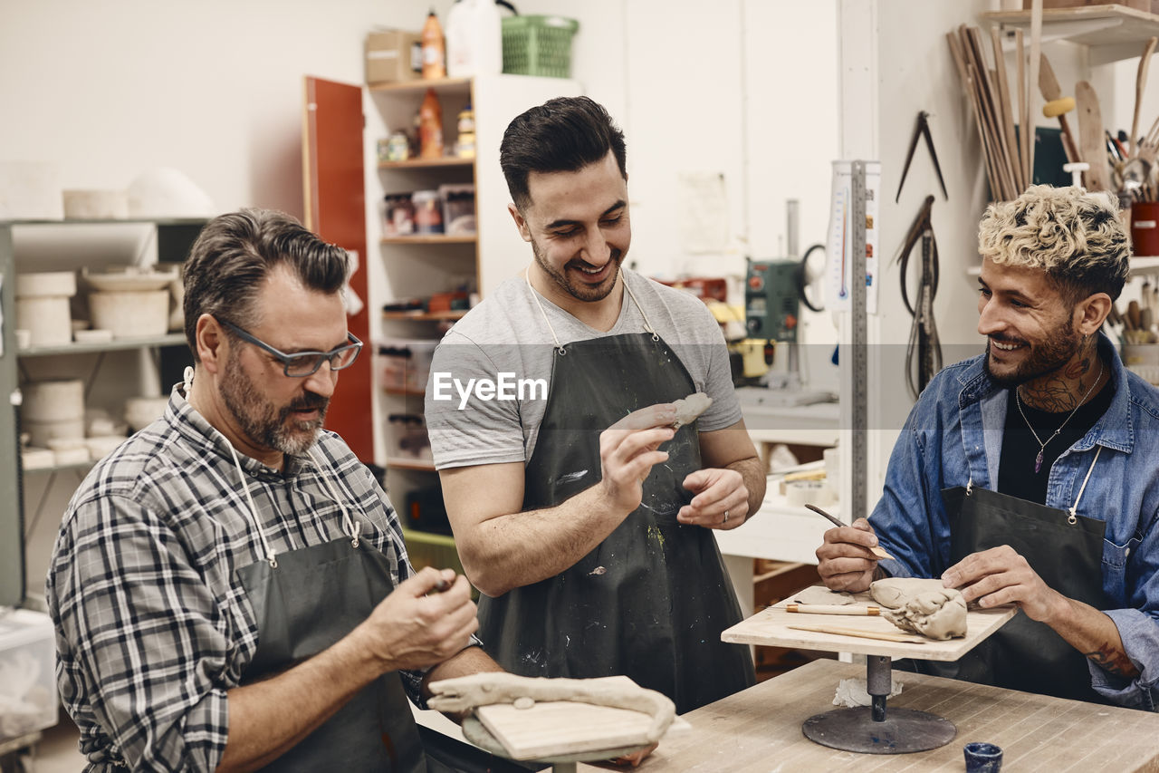 Smiling young man holding molded clay standing by friends sitting with work tool in art class