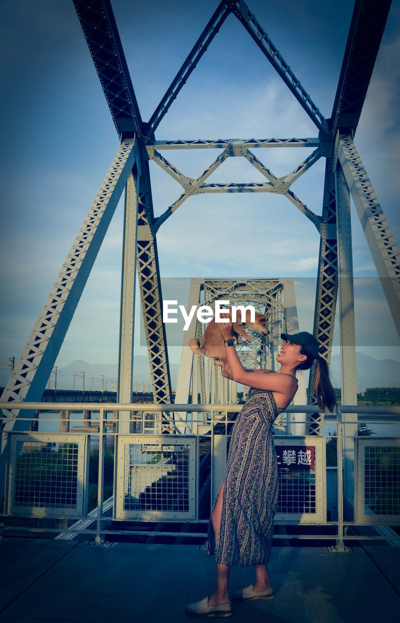 Woman holding dog while standing on bridge against sky