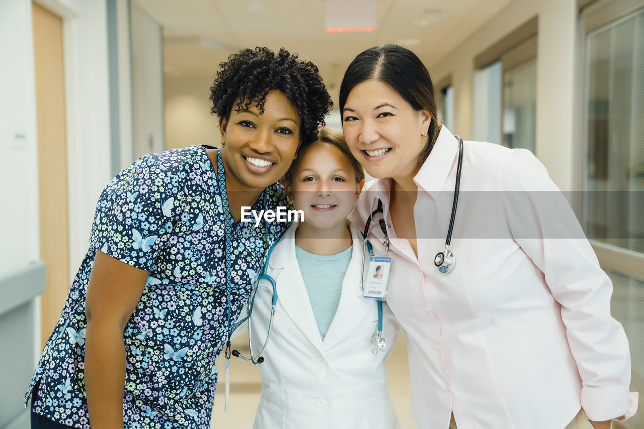 Portrait of confident female doctors with girl wearing lab coat in hospital corridor