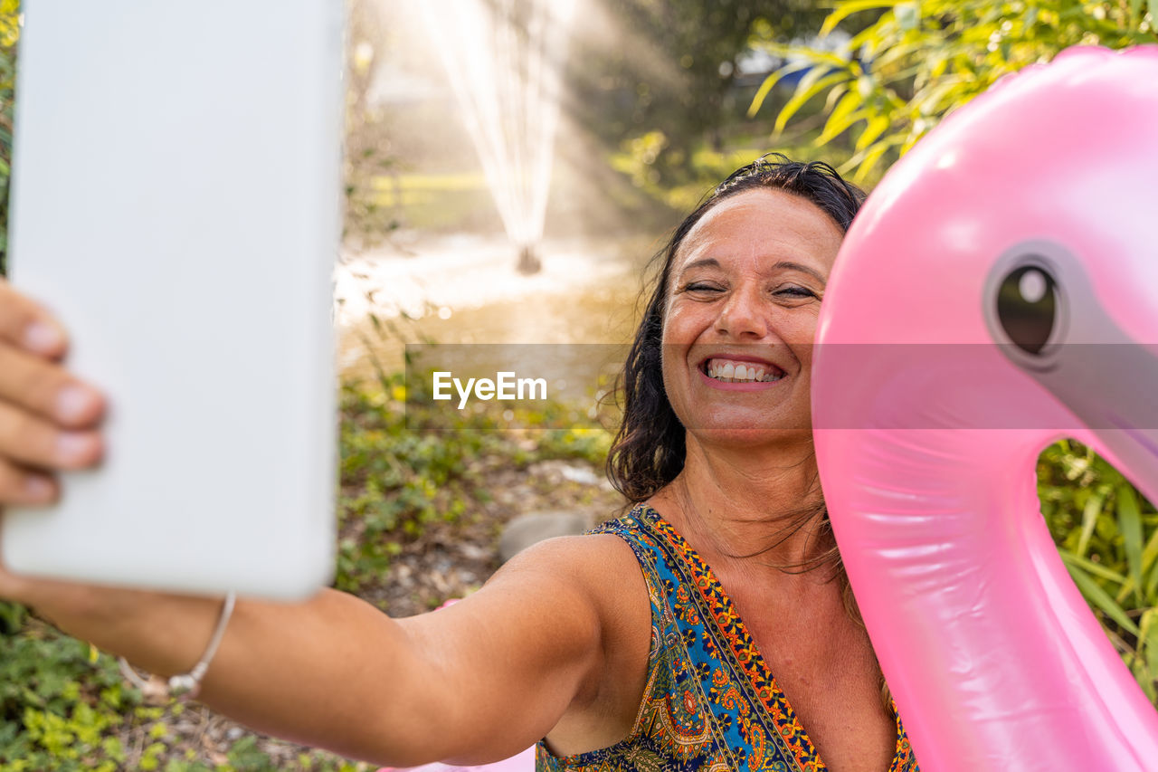 Beautiful smiling middle aged woman taking a selfie sitting on a pink flamingo shaped inflatable toy