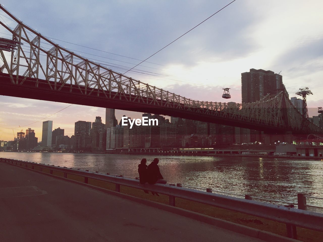 Couple sitting on retaining wall at roosevelt island with queensboro bridge over east river