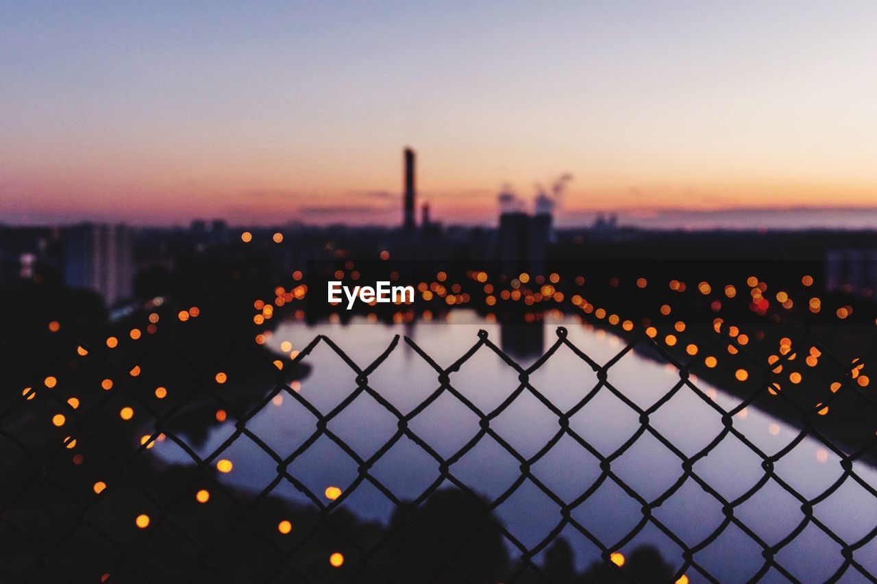 Fence by illuminated city against sky during sunset