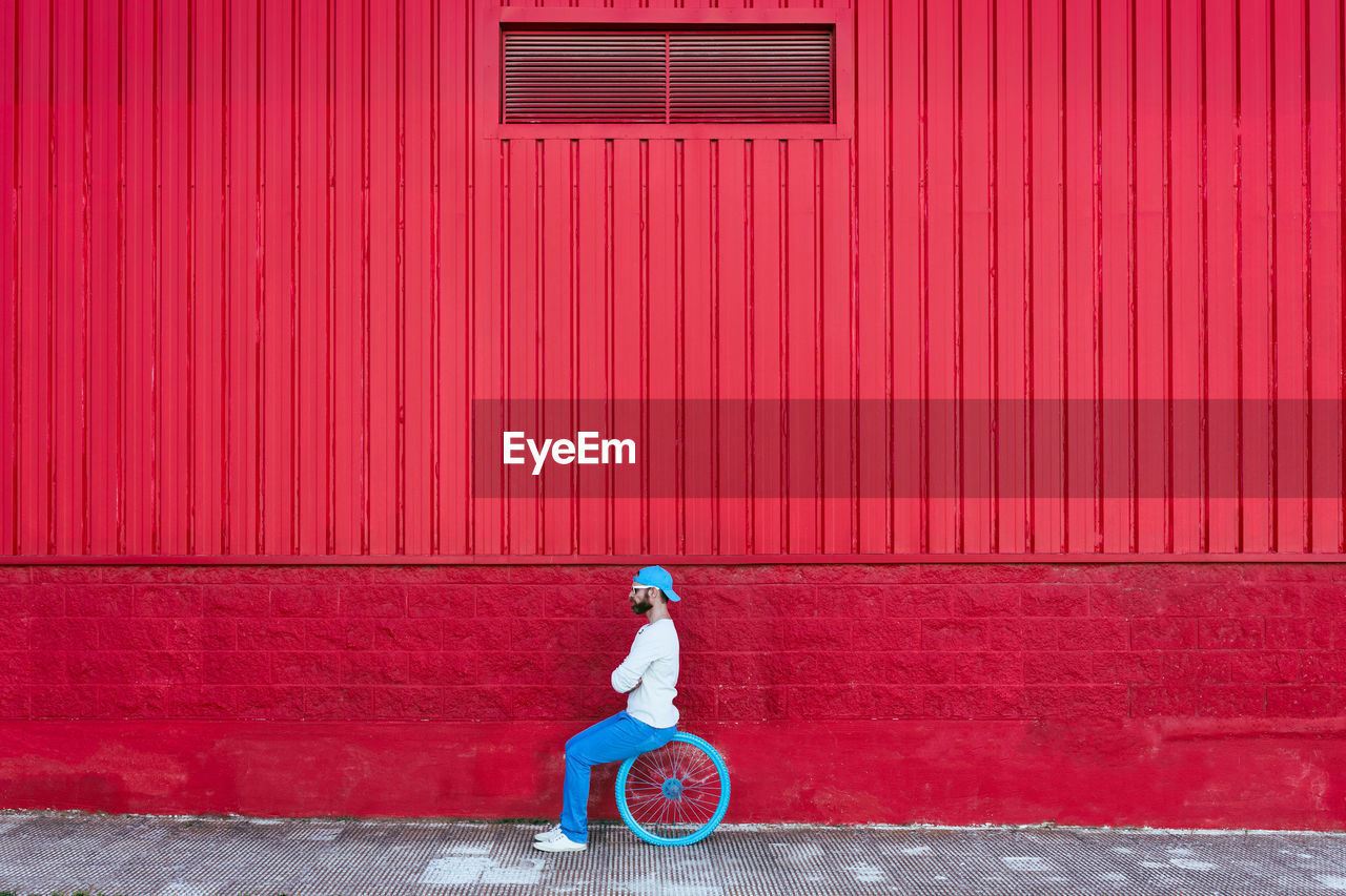 Side view of calm hipster bearded male sitting in bicycle wheel on the street near red background