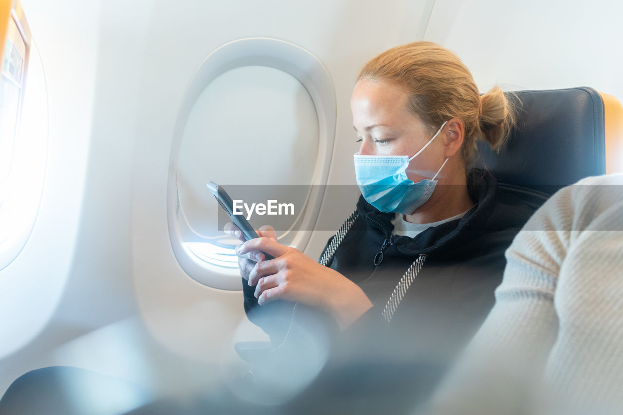 A young woman wearing face mask, using smart phone while traveling on airplane. new normal travel