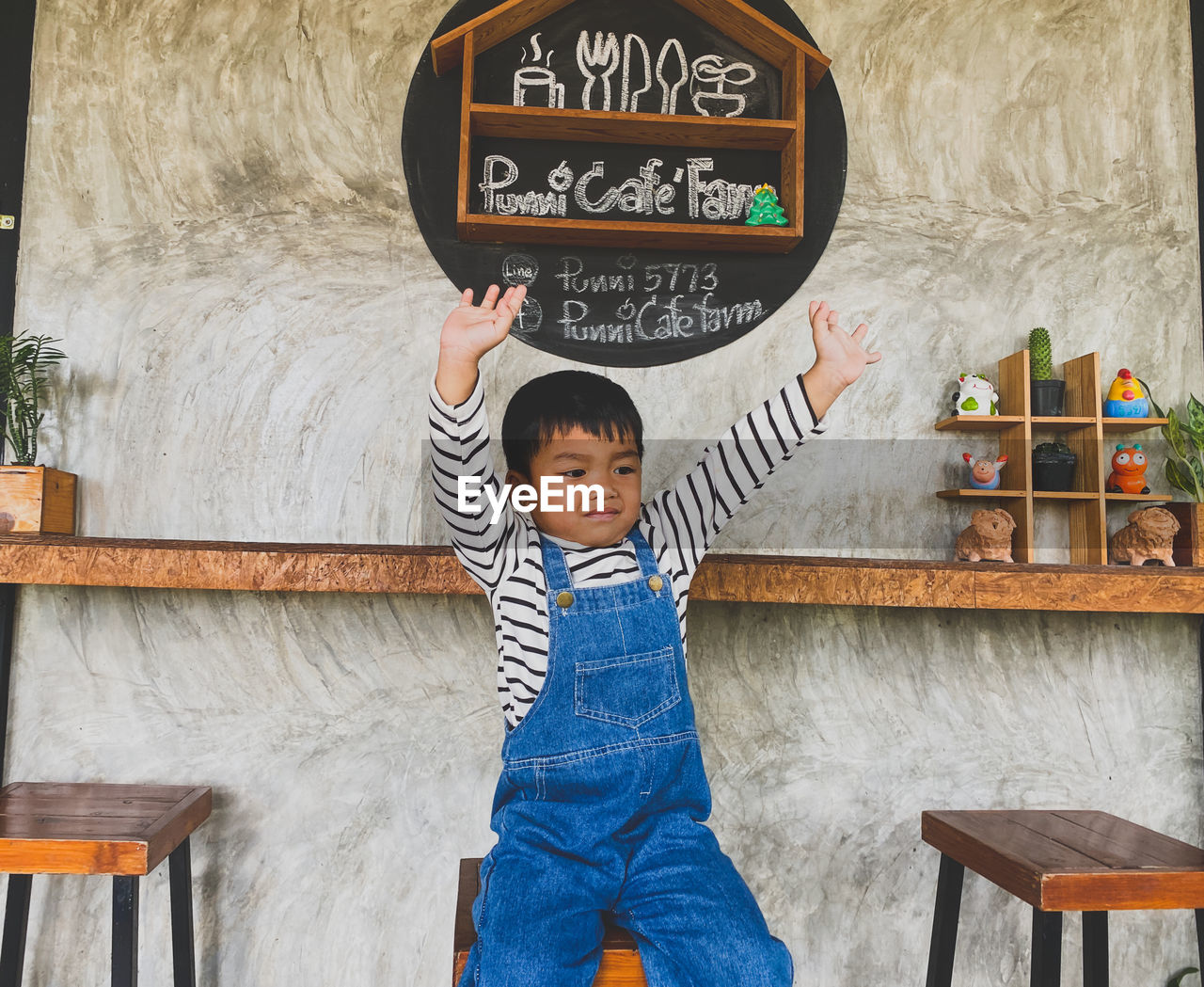 childhood, one person, child, men, casual clothing, furniture, front view, text, standing, three quarter length, wood, full length, cute, board, architecture, innocence, portrait, blackboard, day, indoors, lifestyles, toddler, smiling, happiness, seat, person, communication, emotion, leisure activity, western script, chair, clothing, looking at camera, table