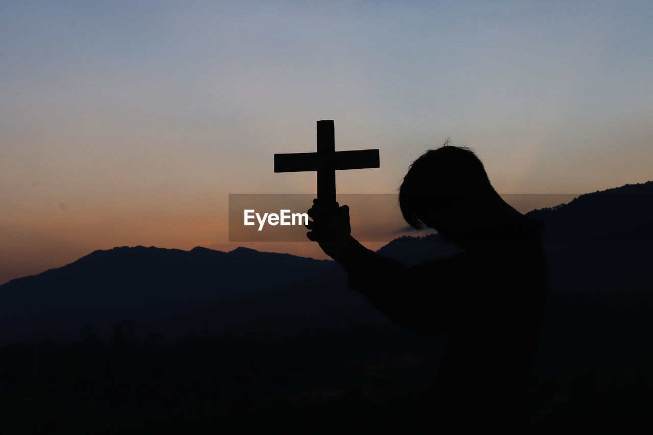 Silhouette person holding cross on mountain against sky during sunset