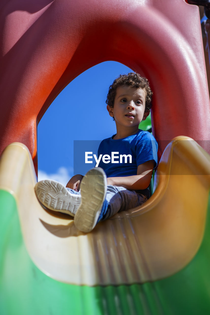 Beautiful caucasian curly haired boy, in playground sitting on top of a slide
