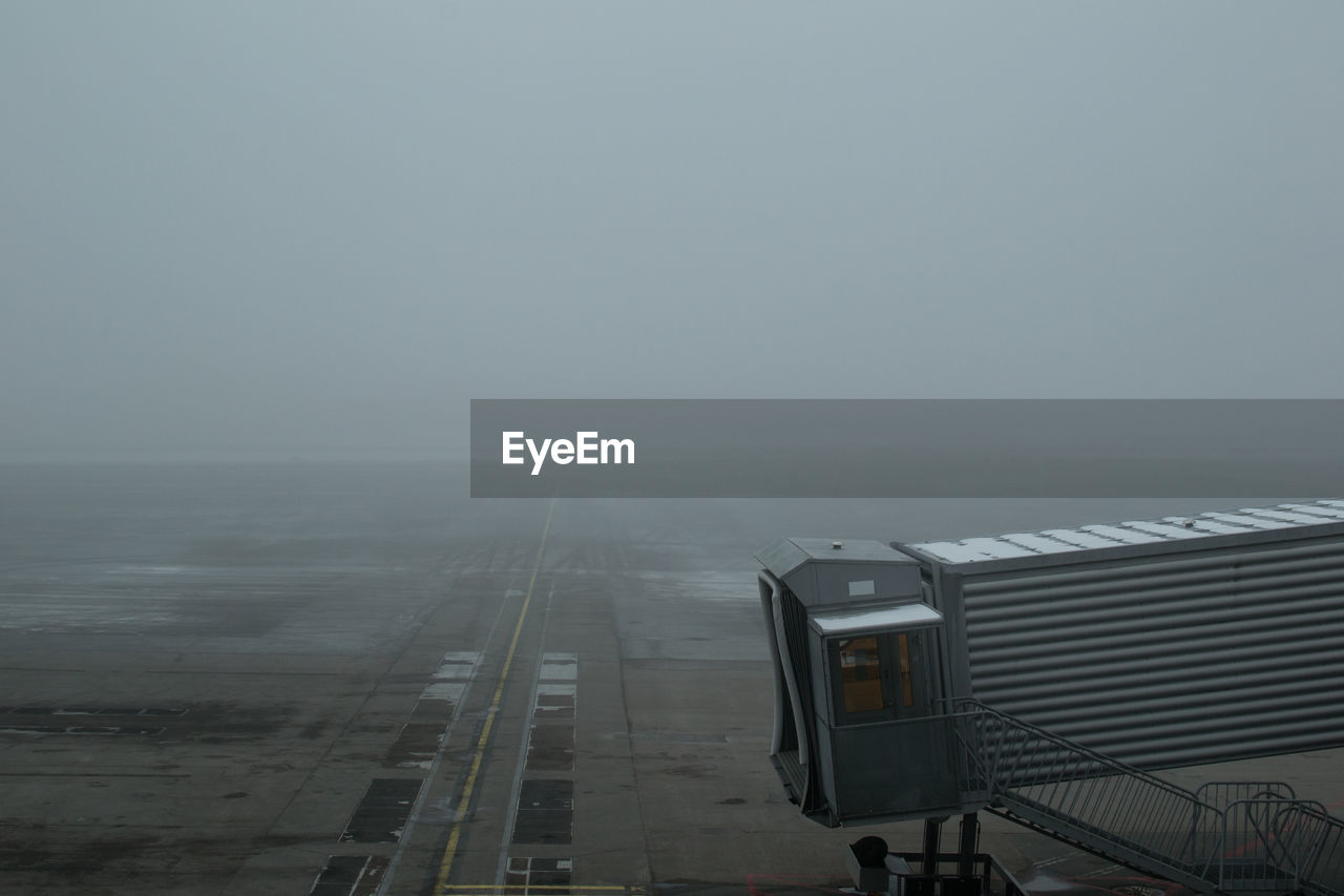 Passenger boarding bridge at airport runway against sky during foggy weather