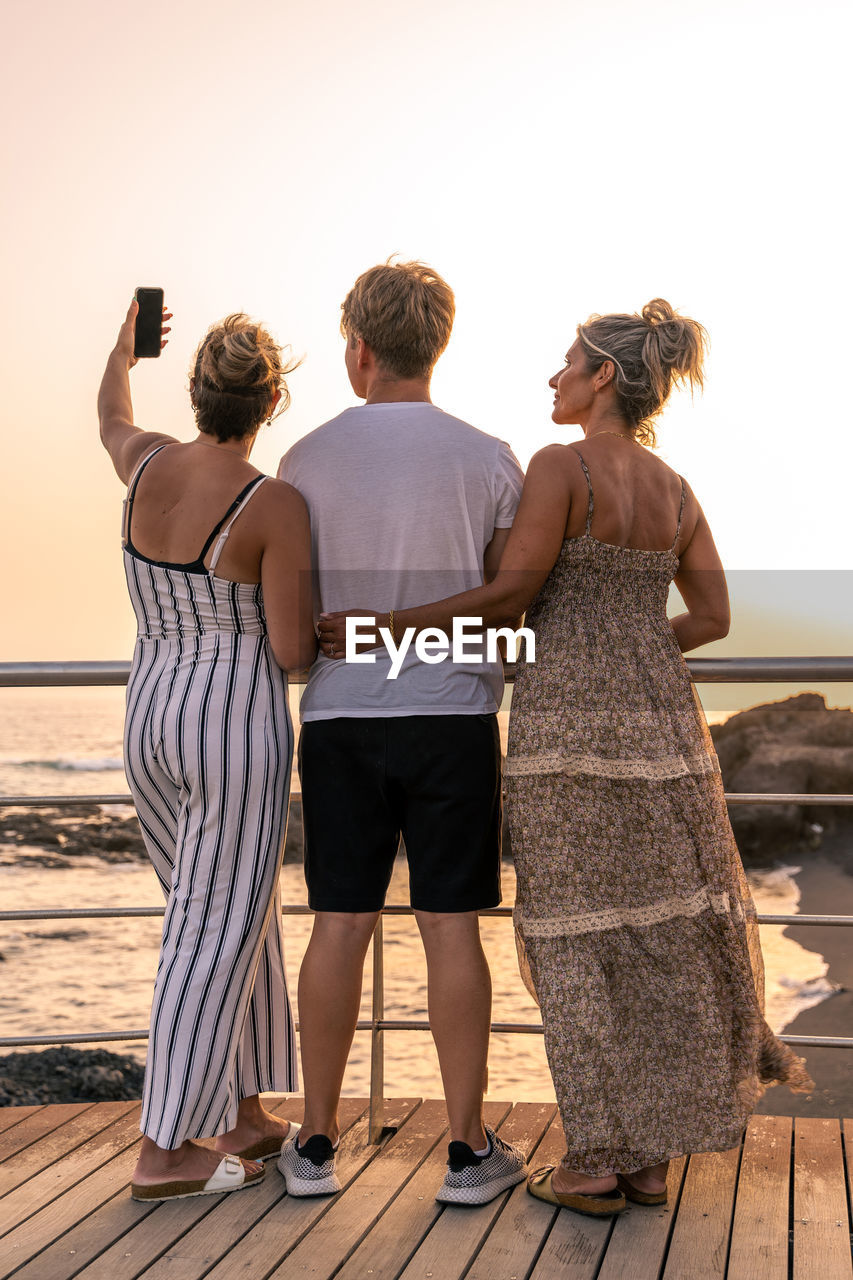 Beautiful mother with her twenty year old sons take a selfie by the sea at sunset - summer concept