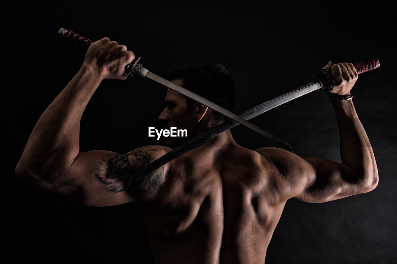 Rear view of shirtless muscular man holding katana swords against black background