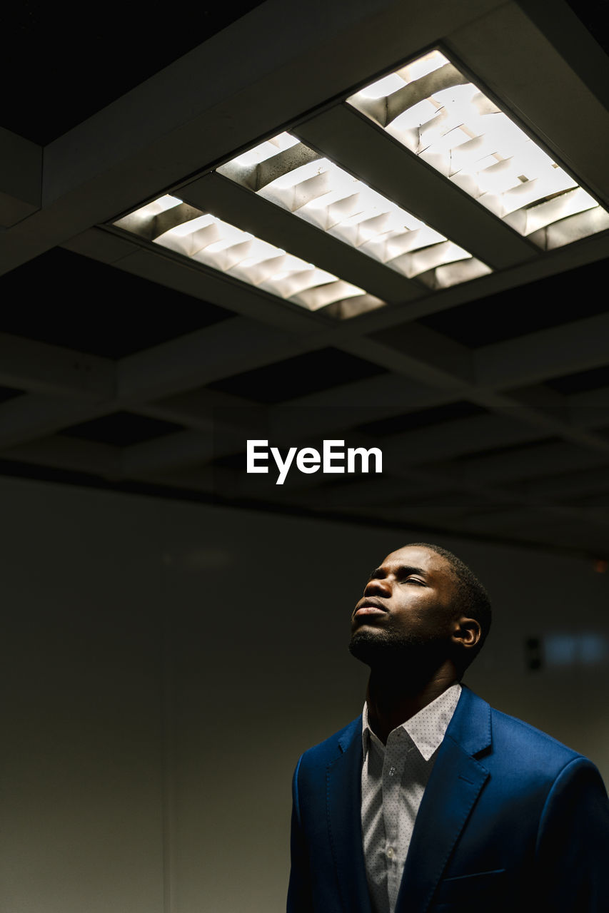 African businessman with eyes closed standing under illuminated fluorescent light in subway