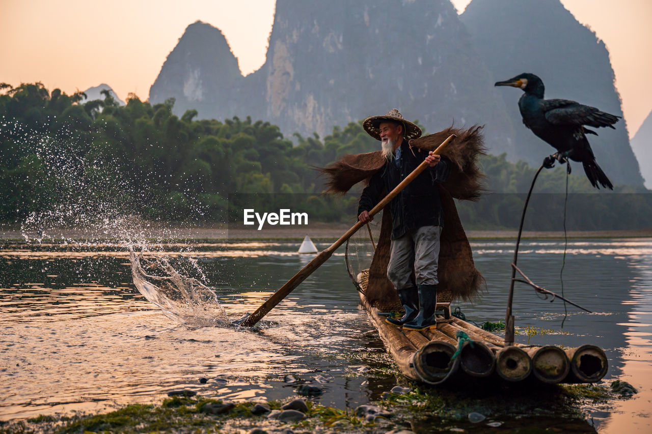 Senior man wearing hat standing on raft against mountain and sky