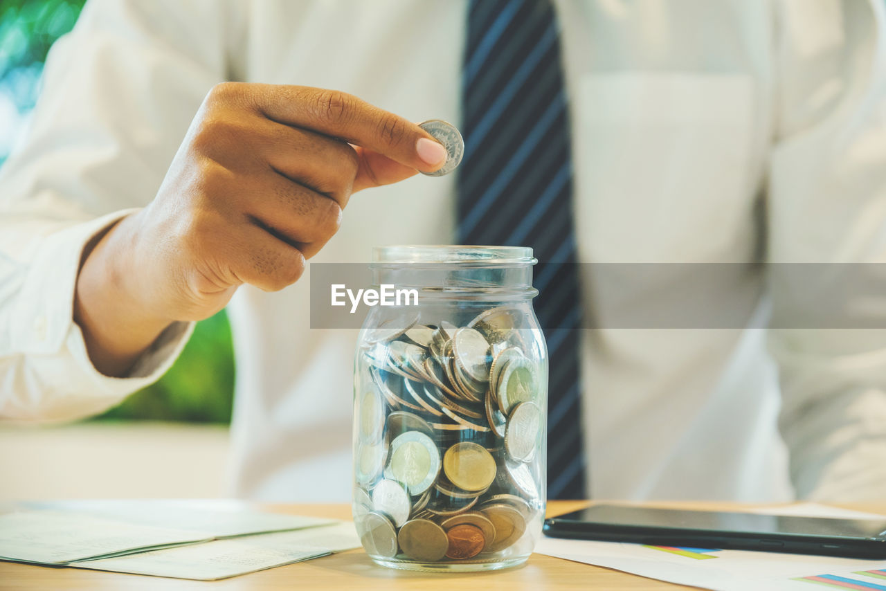 Midsection of man putting coins in glass jar on table