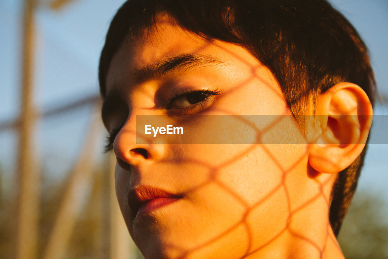 Close-up of boy standing with chain link fence shadow on face outdoors