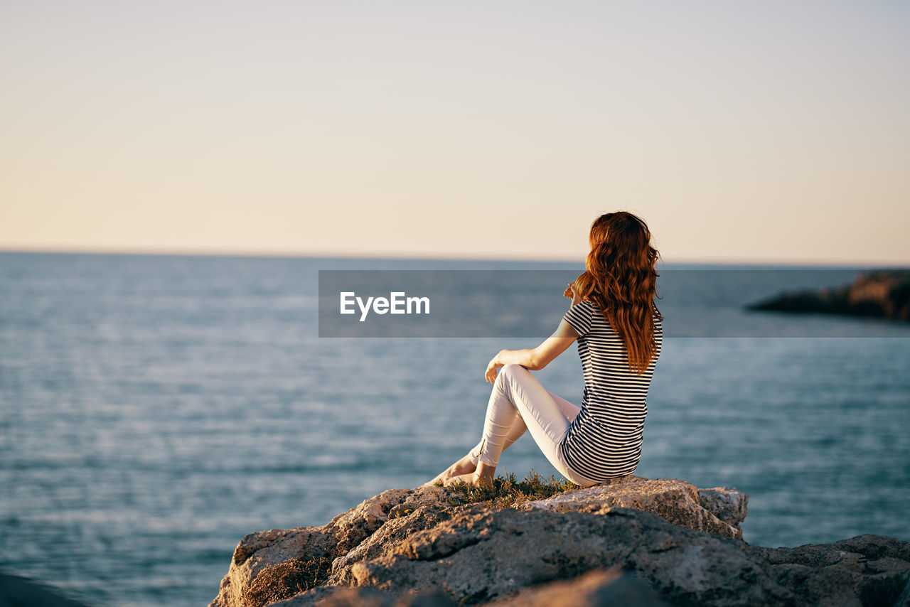 WOMAN ON ROCK BY SEA AGAINST SKY