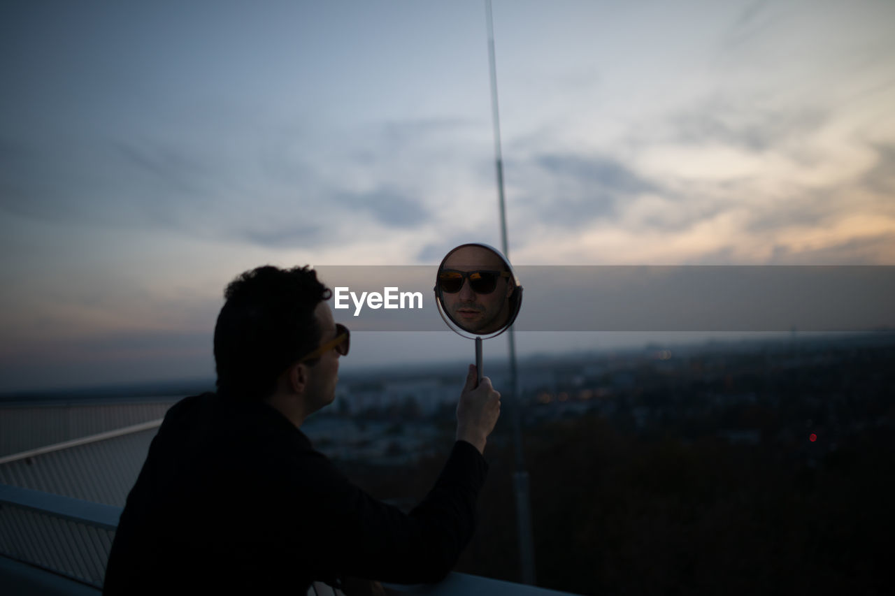 Side view of man holding mirror with his reflection against sky during sunset