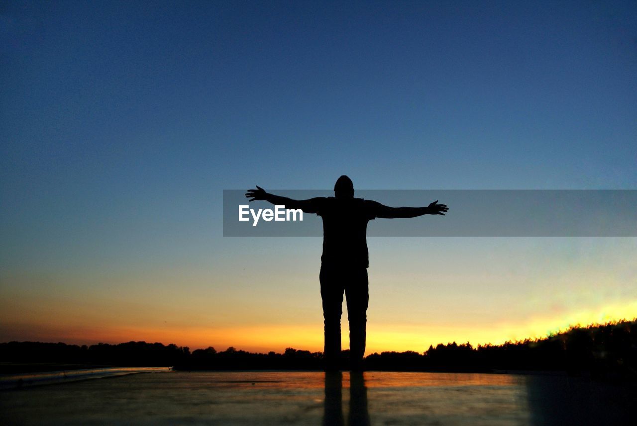 Silhouette man with arms outstretched standing by lake against sky during sunset