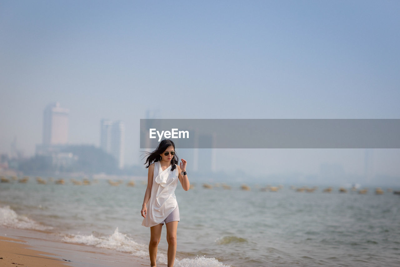 Mid adult woman wearing sunglasses walking at beach against clear sky