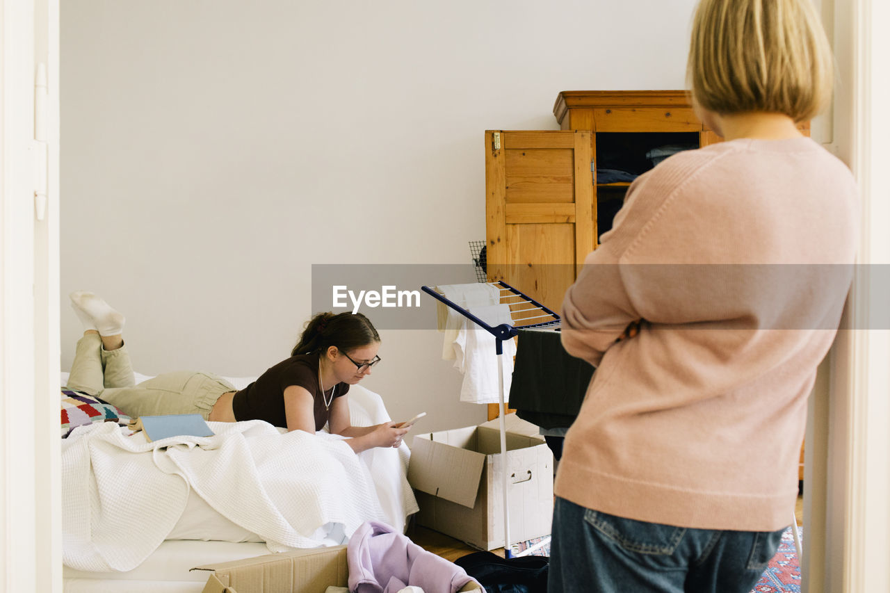 Rear view of parent looking at daughter using smart phone lying on bed at home