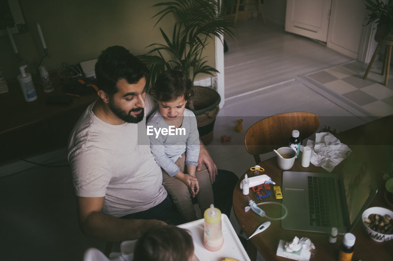 High angle view of father feeding sick son at table in living room