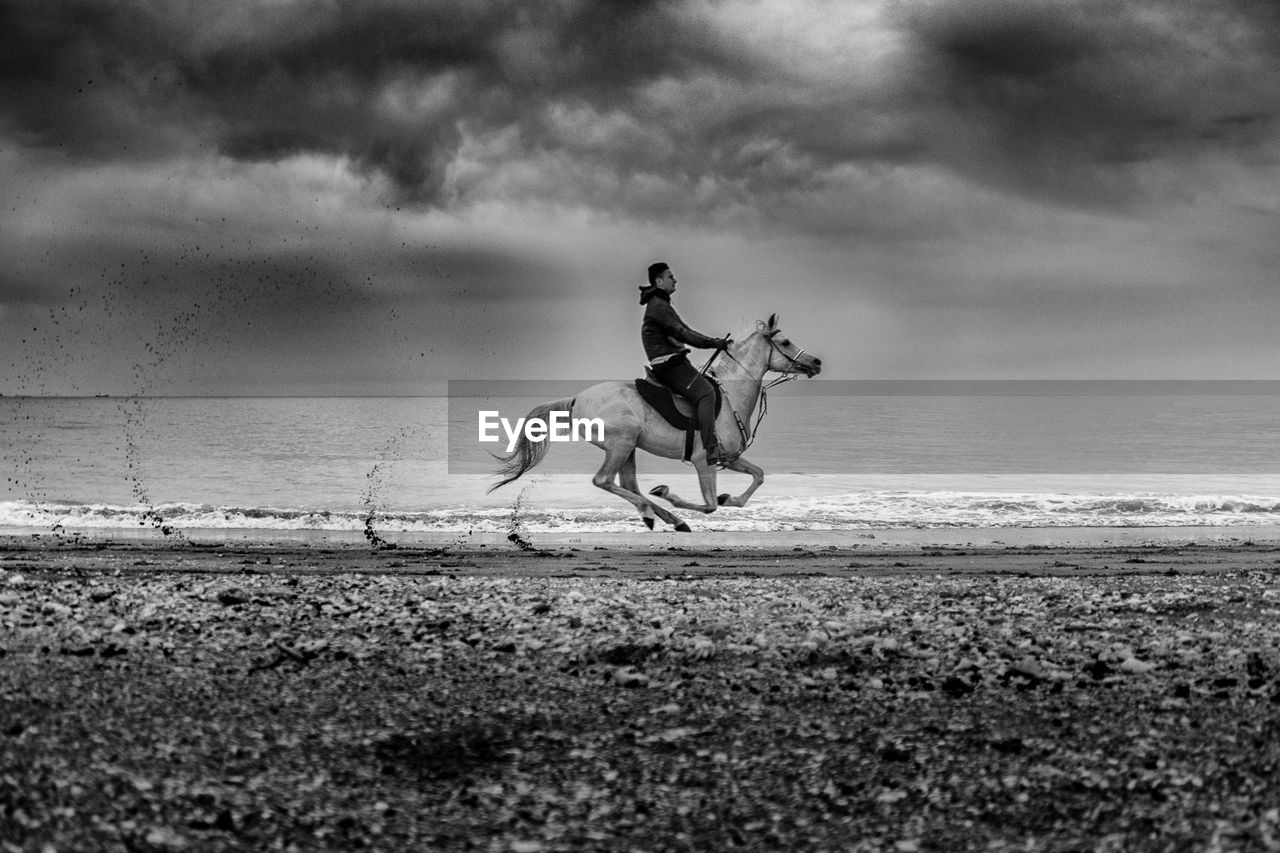 Man riding horse at beach against cloudy sky