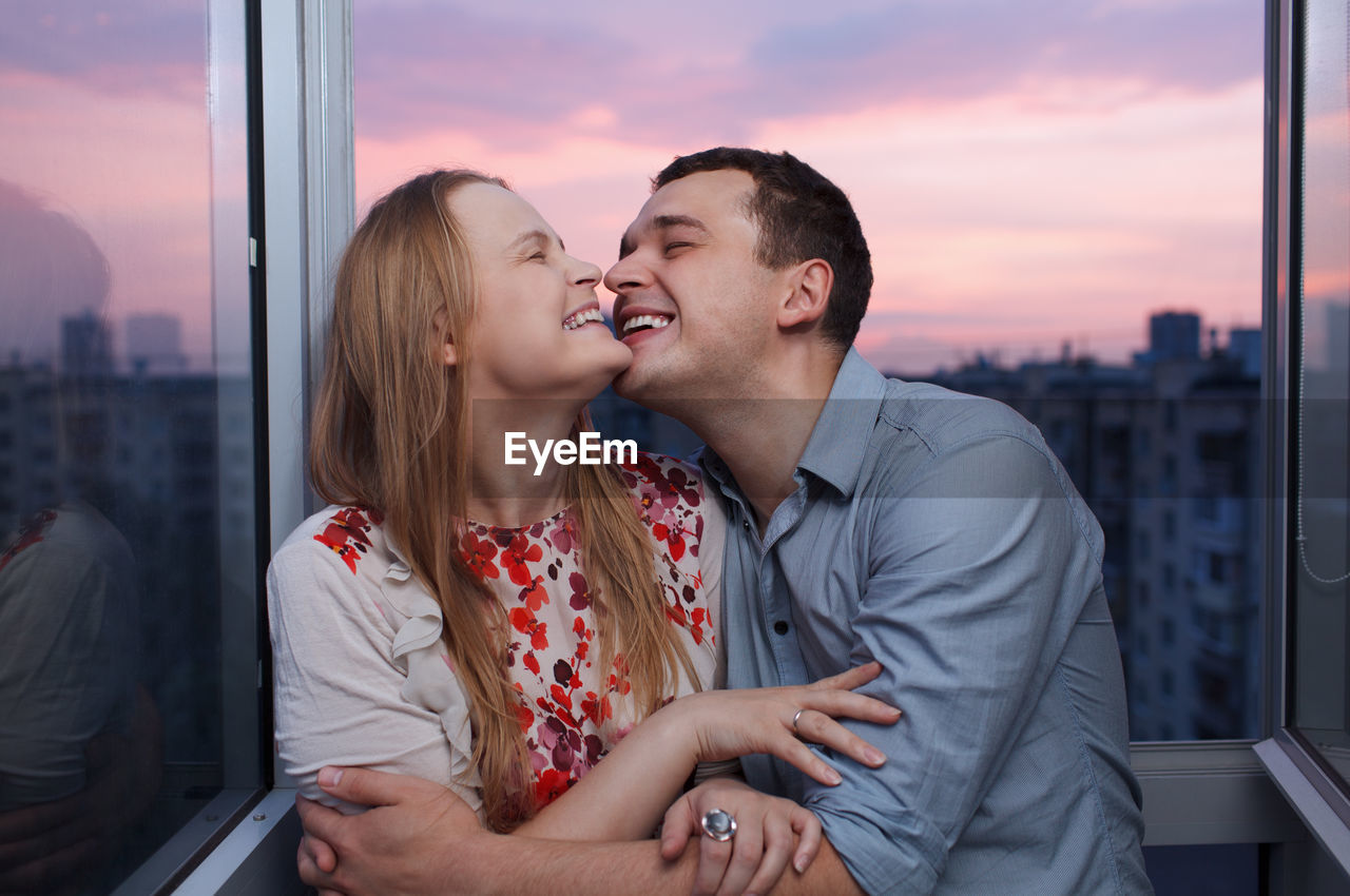 Smiling couple standing at window during sunset