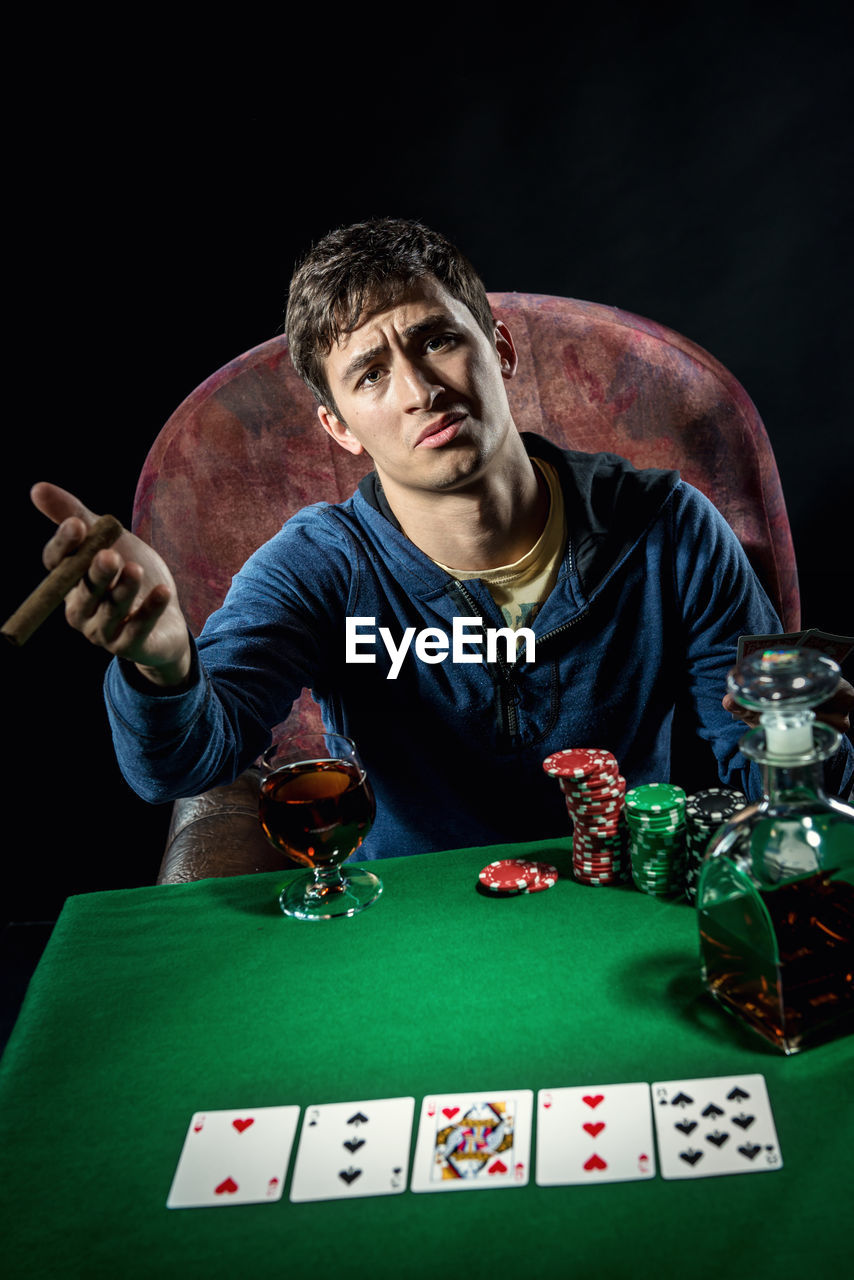Portrait of sad man sitting at poker table with whiskey and cigar against black background