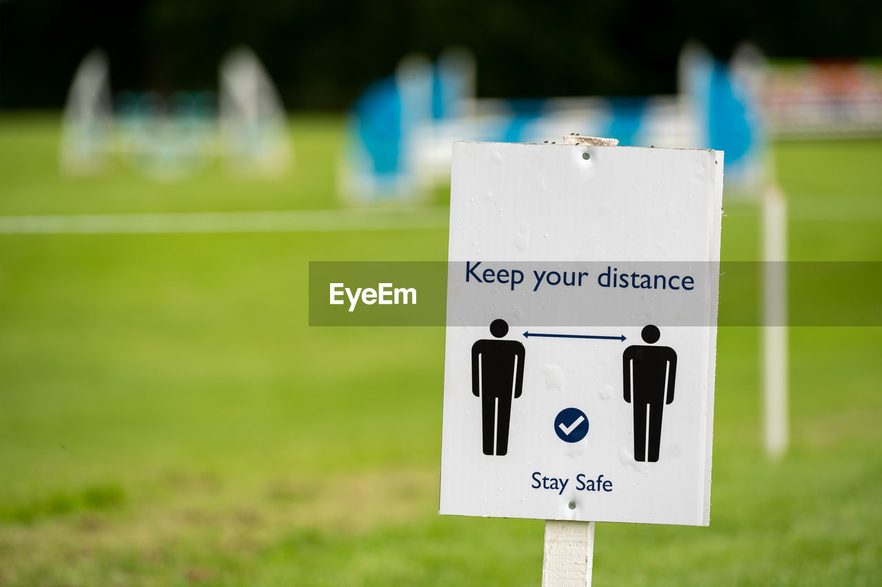 A covid-19 social distancing sign with an out of focus outdoor event in the background.