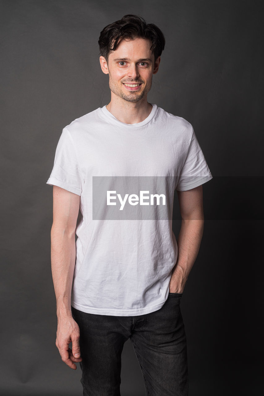 t-shirt, portrait, one person, studio shot, looking at camera, adult, sleeve, casual clothing, men, indoors, white, pocket, standing, beard, smiling, front view, young adult, gray, facial hair, emotion, clothing, photo shoot, happiness, three quarter length, brown hair, hands in pockets, gray background, arm, relaxation, black background, stubble, copy space, person, black, cool attitude, cheerful, trousers, fashion, individuality, lifestyles, hairstyle, jeans